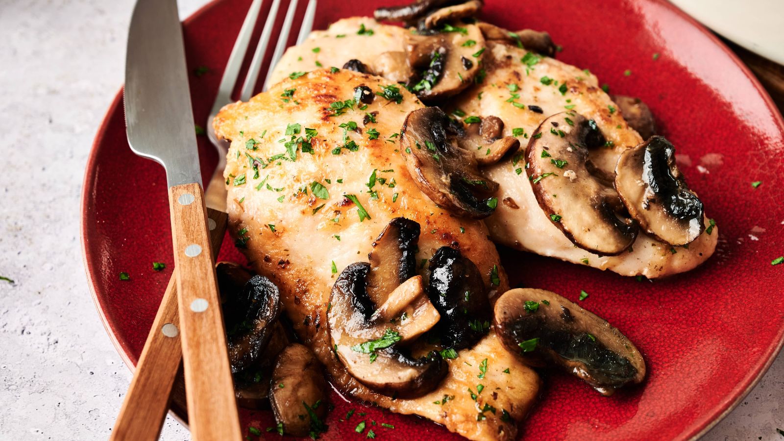 Two pieces of cooked chicken breast topped with sautéed mushrooms and chopped herbs on a red plate, with a fork and knife beside them.