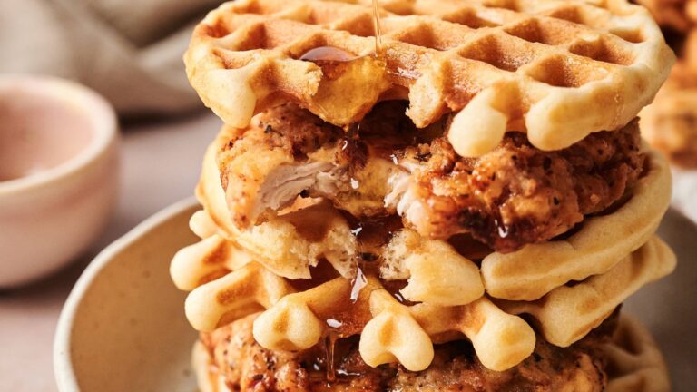 A stack of fried chicken and waffles drizzled with syrup sits on a beige plate, with a small bowl in the background.