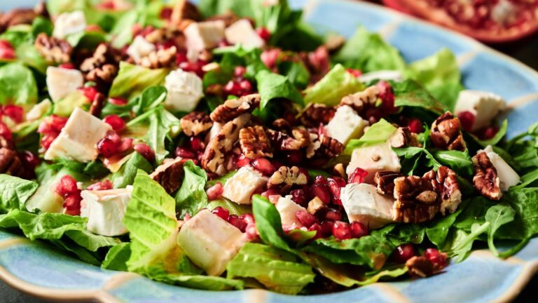 A salad with romaine lettuce, cubed cheese, pomegranate seeds, and pecans is served on a blue plate.