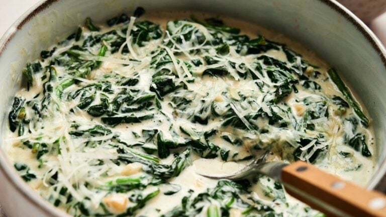 A pan of creamy spinach topped with shredded cheese, with a metal spoon partially visible on the side.