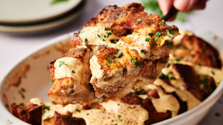 A serving of baked bread pudding topped with creamy sauce and chopped herbs being lifted from a white baking dish.