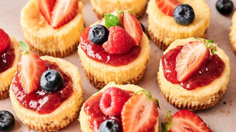 Mini cheesecakes topped with strawberry sauce, fresh strawberries, blueberries, raspberries, and mint leaves arranged on a light surface.