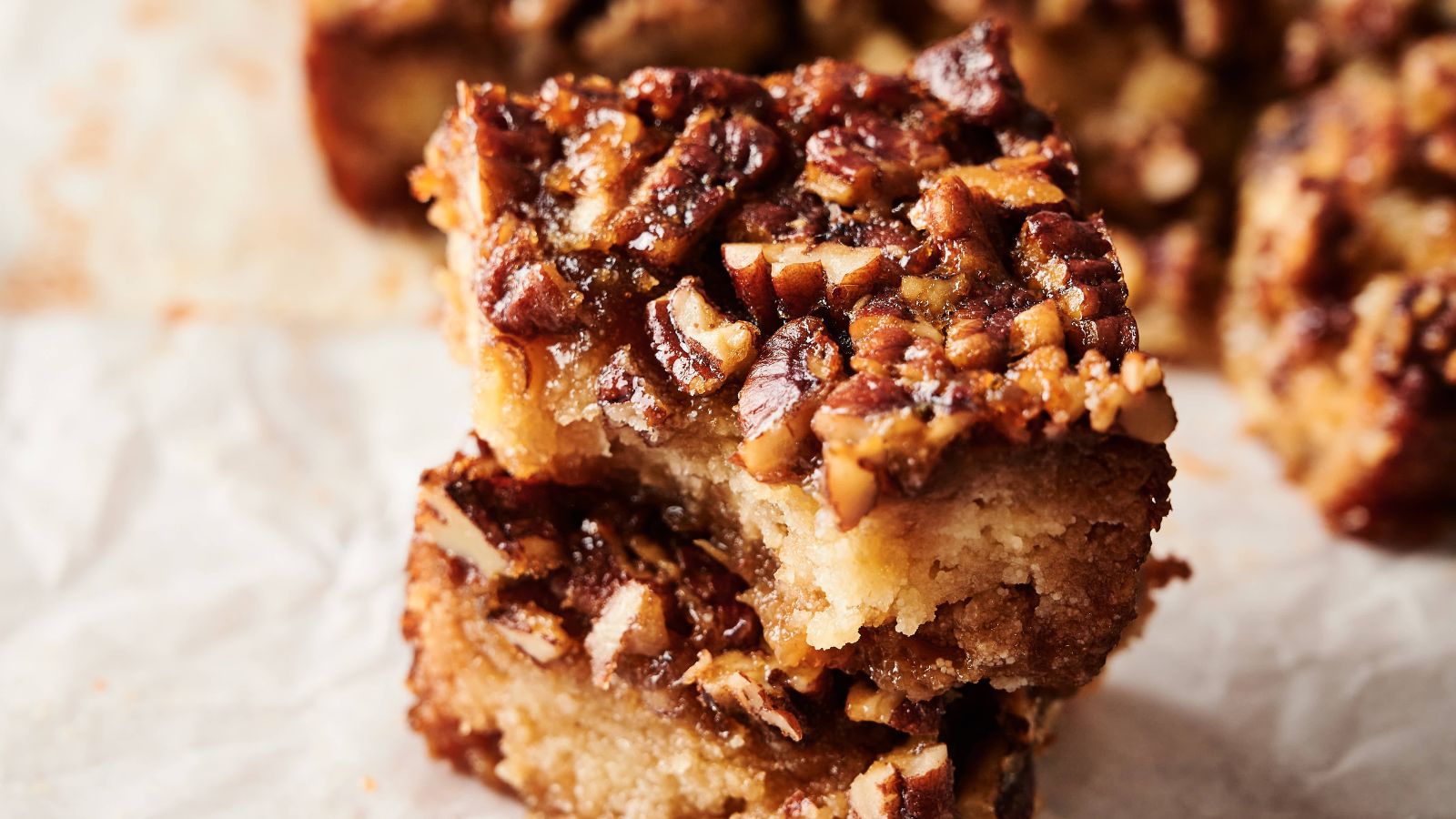 Two pecan-topped dessert bars are stacked on parchment paper, showing a dense, crumbly base and a caramelized nut topping.