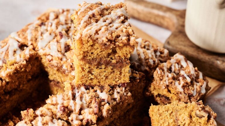 A close-up of several pieces of crumb-topped coffee cake with icing drizzle, stacked on a wooden board next to a mug.