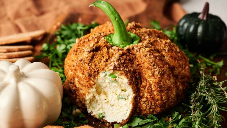 A pumpkin-shaped cheese ball coated in crumbs, with a slice removed to show the creamy interior, surrounded by herbs and decorative gourds.