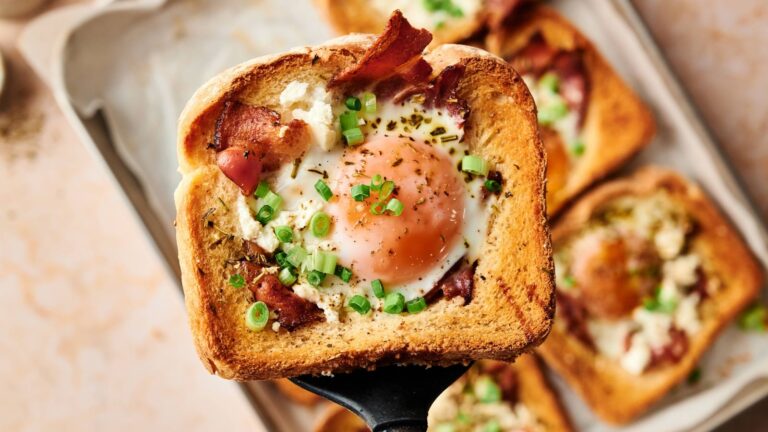A slice of toast with a cooked egg in the center, topped with bacon, green onions, and herbs, held over a tray with more similar toasts in the background.