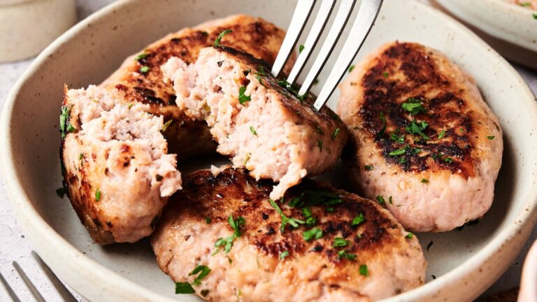 Four browned sausage patties in a bowl, garnished with chopped herbs, with one patty being cut open with a fork to show its inside.
