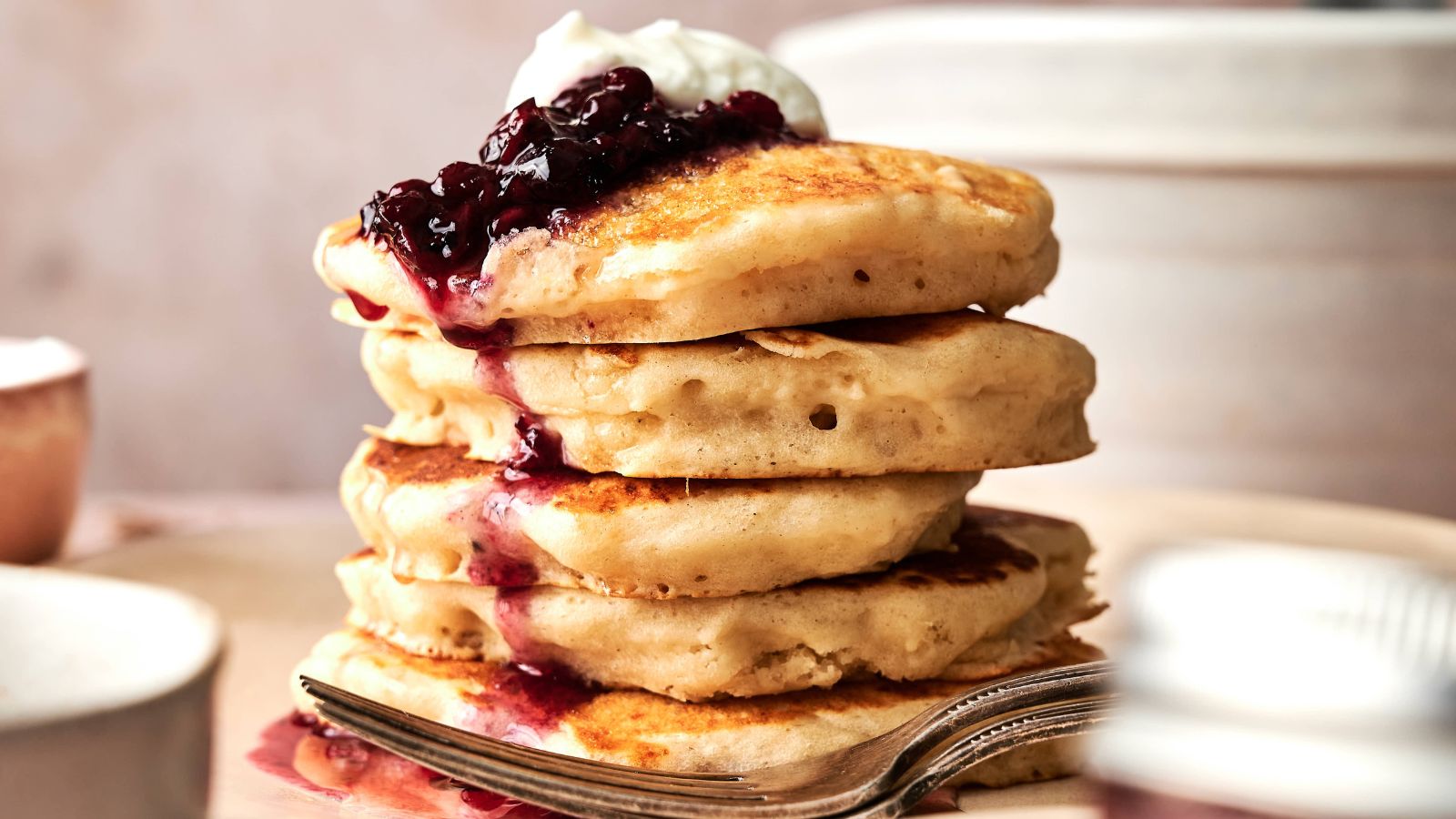 A stack of four pancakes topped with berry compote and a dollop of cream, served on a plate with a fork.