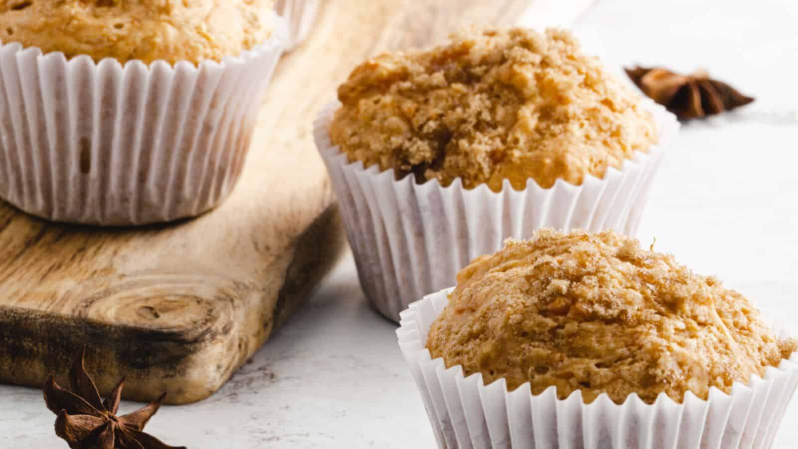 Three muffins in white paper liners are displayed on a wooden board and a white surface, with a star anise in the foreground.