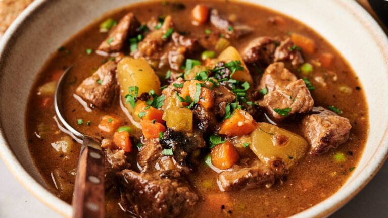 A bowl of beef stew with chunks of beef, potatoes, carrots, and peas in a brown broth, garnished with chopped parsley.