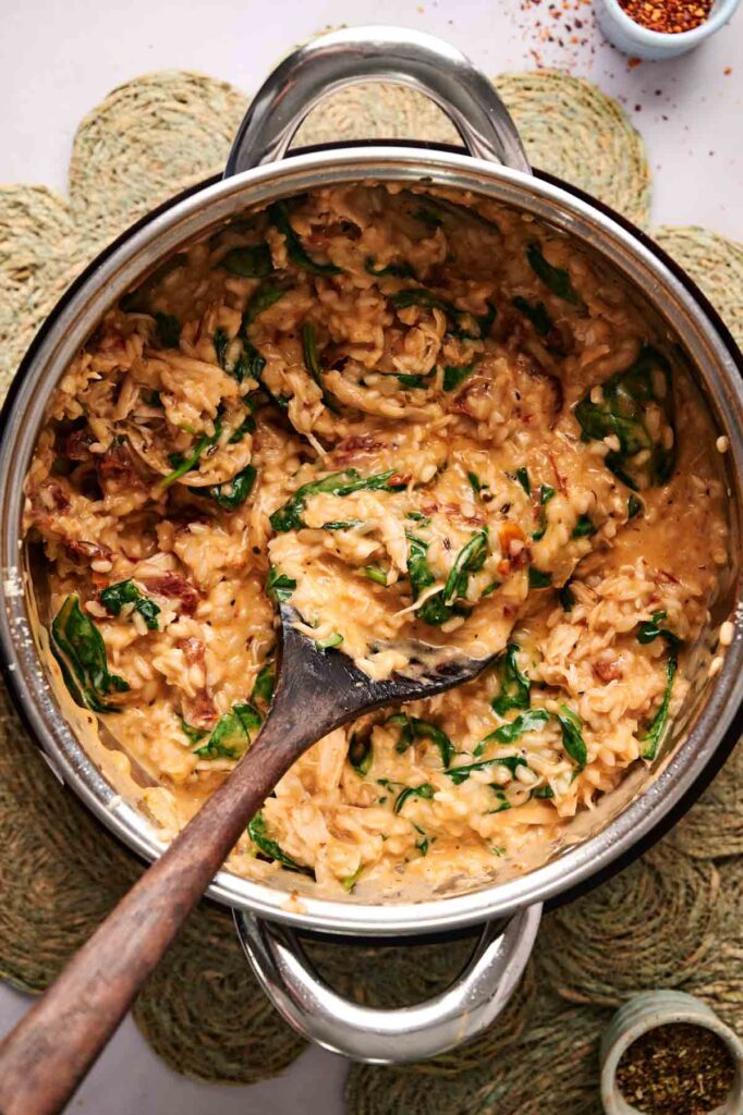 A pot of creamy Marry Me Chicken Risotto with spinach and sun-dried tomatoes, stirred with a wooden spoon, sits on a woven mat beside spices.