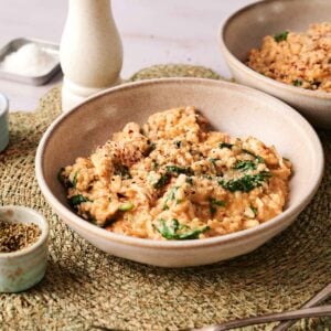 A bowl of creamy Marry Me Chicken Risotto with spinach and black pepper, placed on a woven placemat with a pepper grinder, small dishes of seasoning, and two forks nearby.