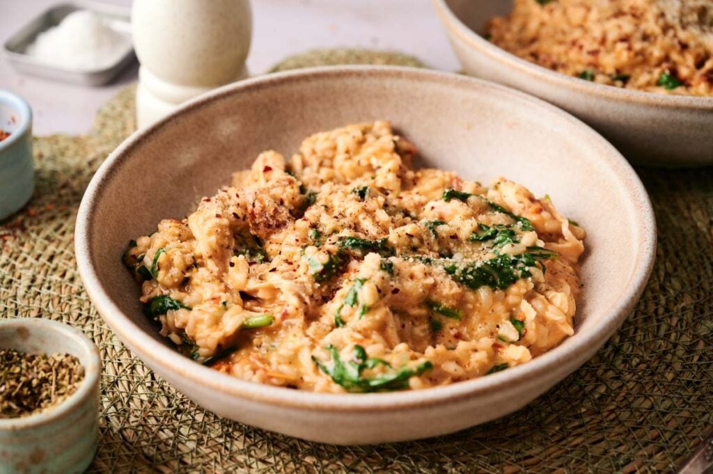 A bowl filled with creamy Marry Me Chicken Risotto, mixed with spinach and topped with grated cheese and black pepper, sits on a woven mat.