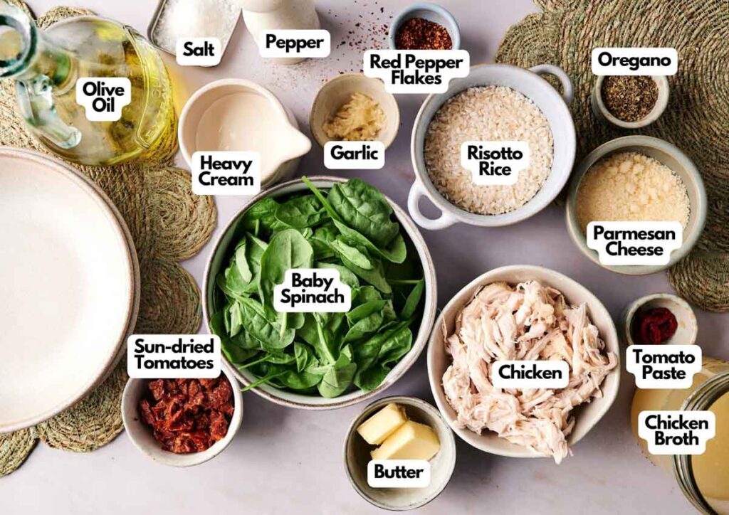 Various Marry Me Chicken Risotto ingredients are arranged on a table, including baby spinach, chicken, risotto rice, sun-dried tomatoes, seasonings, cheese, butter, broth, garlic, and olive oil.