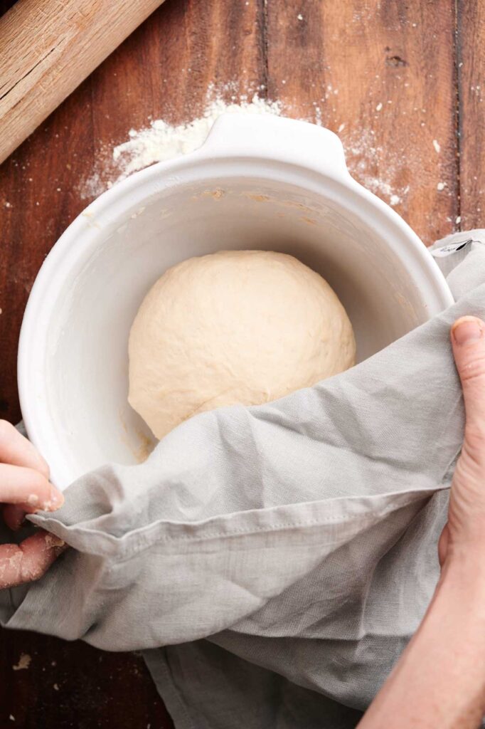 A round ball of pita bread dough in a white bowl is partially covered with a gray cloth on a wooden surface sprinkled with flour.