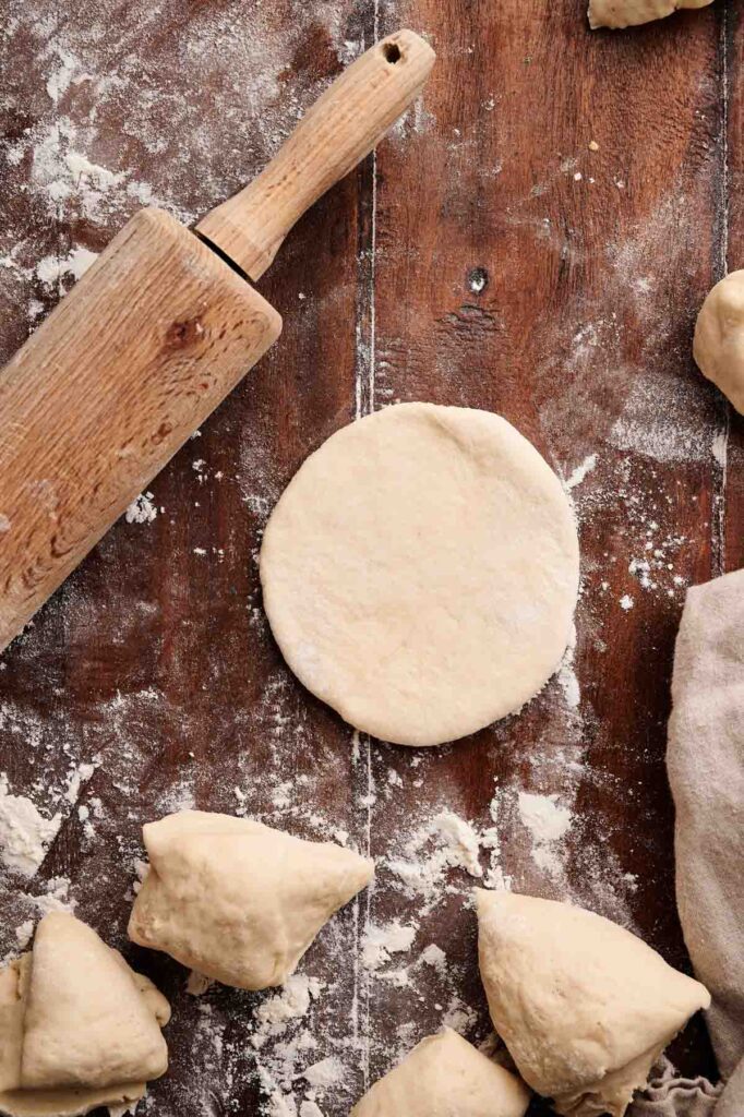 Round dough, dough pieces, and a wooden rolling pin on a floured wooden surface, suggesting pita bread or pizza preparation.