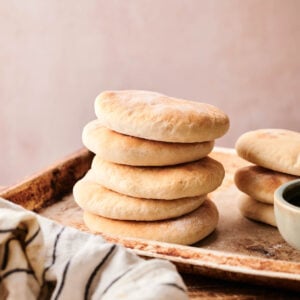 A stack of round, golden Pita Bread sits on a wooden tray next to a striped cloth and a small ceramic bowl.