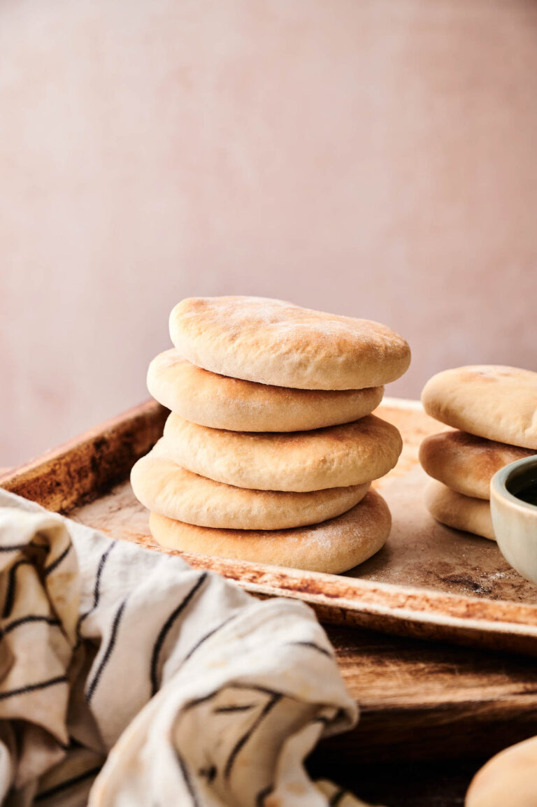 A stack of round, golden Pita Bread sits on a wooden tray next to a striped cloth and a small ceramic bowl.