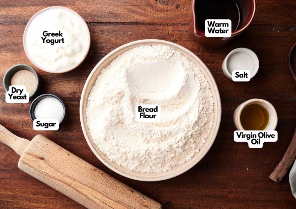 A bowl of bread flour for Pita Bread is surrounded by labeled ingredients: Greek yogurt, dry yeast, sugar, warm water, salt, and virgin olive oil, with a rolling pin resting on a wooden table.