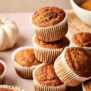 Pumpkin Muffins in paper liners are stacked and arranged on a light surface, with a white pumpkin and a bowl in the background.