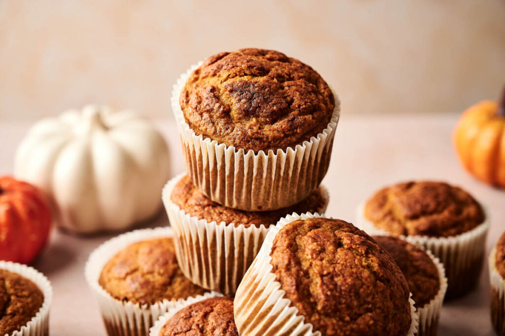 A stack of pumpkin muffins in paper liners sits on a light surface, with small decorative pumpkins in the background, creating a cozy autumn scene perfect for showcasing delicious Pumpkin Muffins.