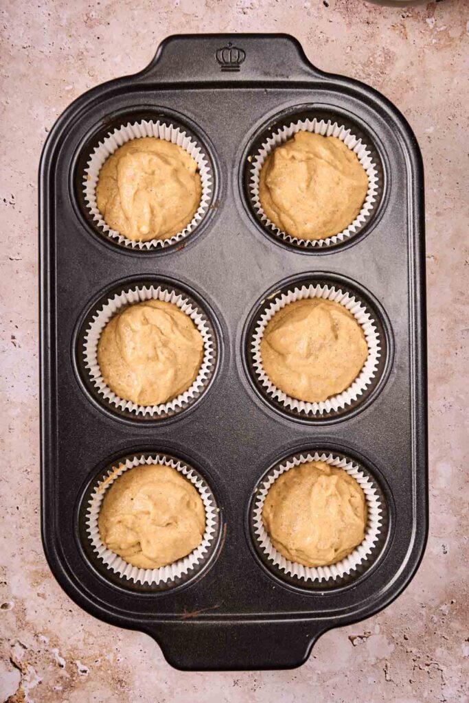 A six-cup muffin pan with paper liners, each filled with pumpkin muffins batter, placed on a textured countertop.