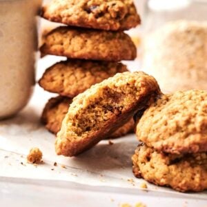 A stack of Pumpkin Oatmeal Cookies, with one cookie broken in half, rests on parchment paper beside a cup and a glass jar in the background.