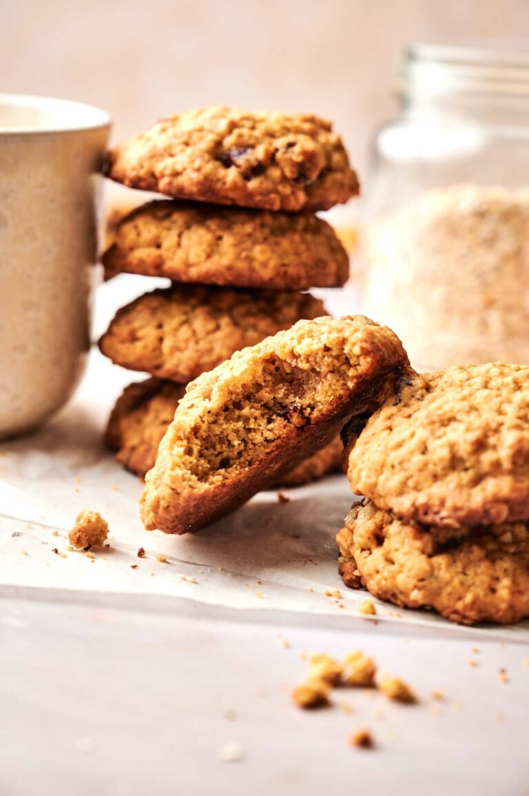 A stack of Pumpkin Oatmeal Cookies, with one cookie broken in half, rests on parchment paper beside a cup and a glass jar in the background.