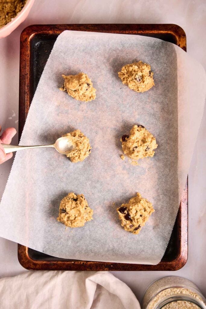 A baking tray lined with parchment paper holds six scoops of Pumpkin Oatmeal Cookie dough, with a hand using a spoon to place another scoop.