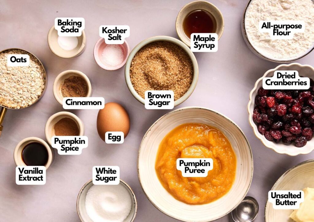 A variety of baking ingredients for Pumpkin Oatmeal Cookies&mdash;oats, pumpkin puree, brown sugar, dried cranberries, flour, an egg, and spices&mdash;are arranged in bowls on a light surface with labels.