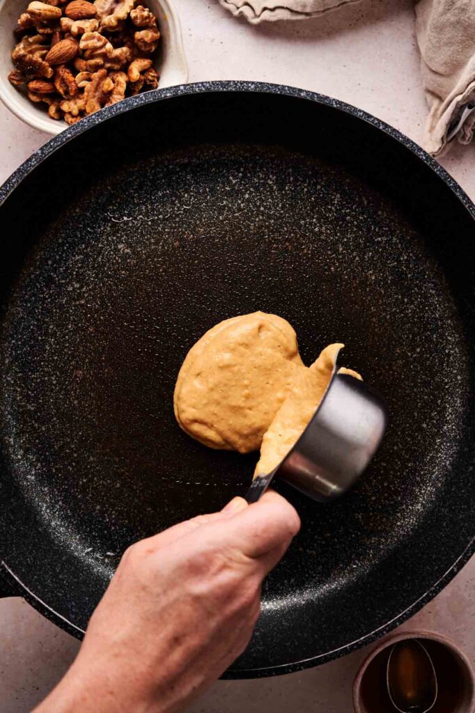 A hand pours pumpkin pancake batter from a measuring cup onto a black frying pan, with a bowl of walnuts visible nearby.