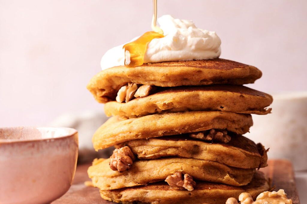 A stack of pumpkin pancakes topped with whipped cream and syrup, with walnuts between the layers, sits on a wooden surface next to a ceramic bowl.