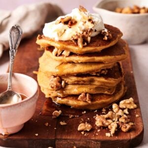 A stack of pumpkin pancakes topped with whipped cream and chopped walnuts sits on a wooden board, with extra walnuts and a small bowl with a spoon nearby.