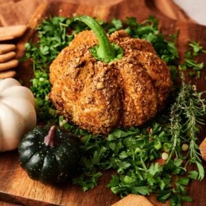 A Pumpkin-Shaped Cheeseball coated in seasoning sits on chopped parsley, surrounded by crackers, herbs, and small decorative gourds on a wooden board.