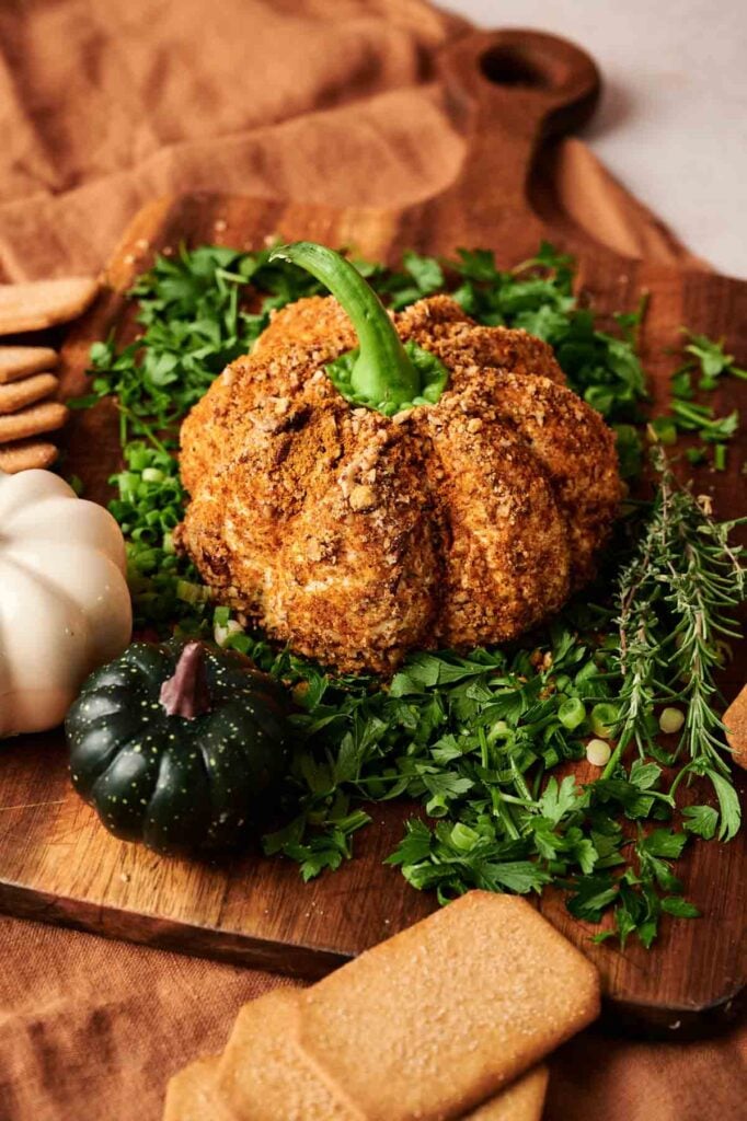 A Pumpkin-Shaped Cheeseball coated in seasoning sits on chopped parsley, surrounded by crackers, herbs, and small decorative gourds on a wooden board.