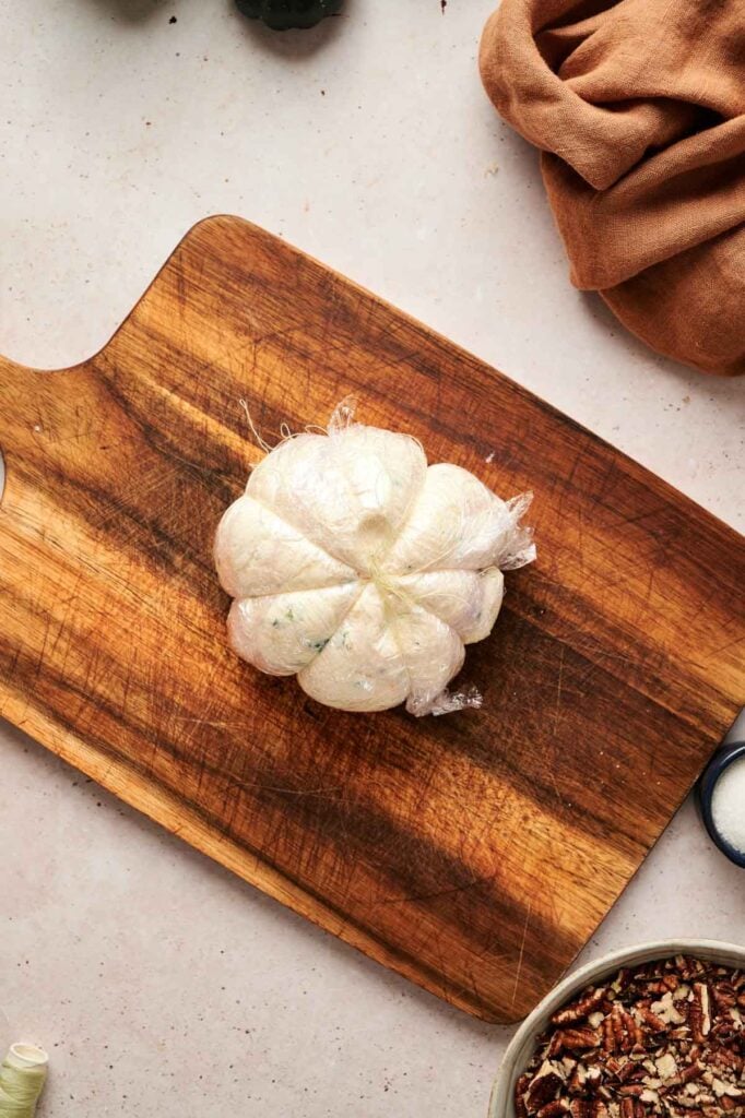A Pumpkin-Shaped Cheeseball wrapped in plastic wrap and tied with string sits on a wooden cutting board, surrounded by kitchen items.