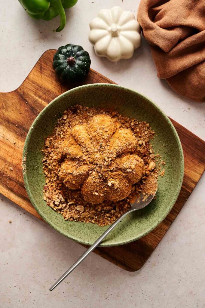 A Pumpkin-Shaped Cheeseball sits in a green bowl filled with crumbs on a wooden board, surrounded by decorative pumpkins and a brown cloth for a festive touch.