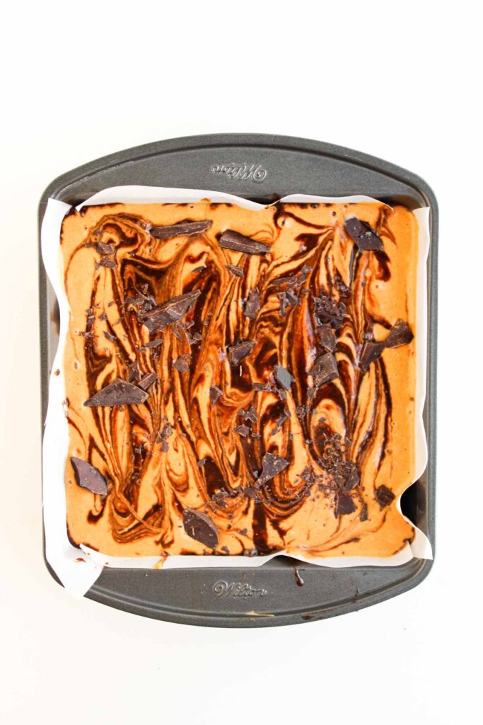 A square baking pan lined with parchment paper filled with swirled Pumpkin Spice Fudgy Brownies batter, topped with chunks of chocolate and creamy peanut butter for a decadent treat.