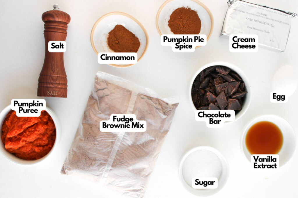 Various baking ingredients labeled on a white surface, ready to make Pumpkin Spice Fudgy Brownies&mdash;featuring salt, cinnamon, pumpkin puree, fudge brownie mix, pumpkin pie spice, chocolate bar, sugar, vanilla extract, egg, and cream cheese.