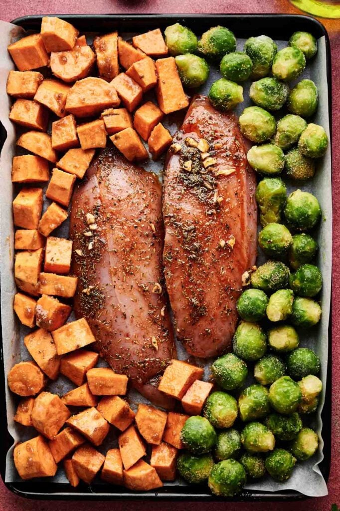 Raw seasoned chicken breasts, cubed sweet potatoes, and Brussels sprouts arranged on a parchment-lined baking sheet&mdash;perfect for an easy Sheet Pan Thanksgiving Dinner.