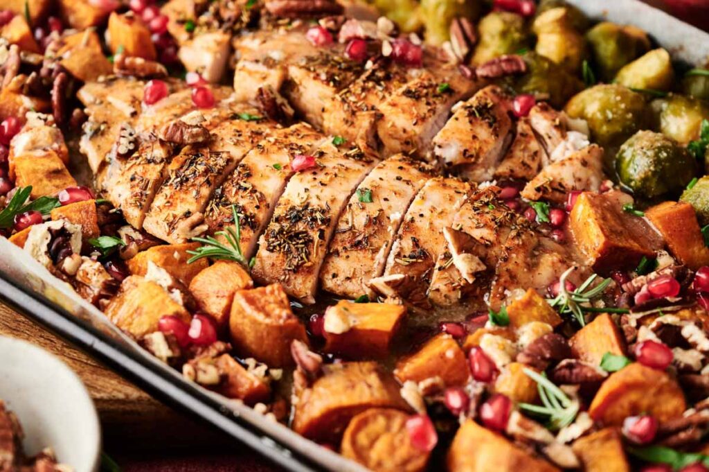 Sliced roasted chicken breast on a sheet pan with sweet potatoes, Brussels sprouts, pecans, rosemary, and pomegranate seeds&mdash;perfect for an easy Sheet Pan Thanksgiving Dinner.