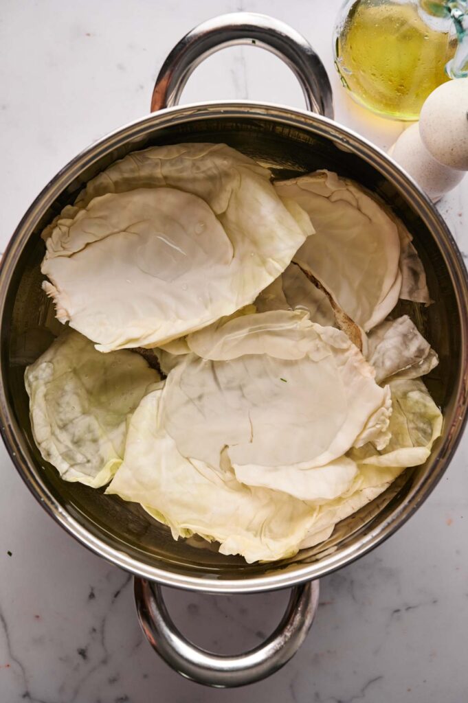 A stainless steel pot filled with large, pale green cabbage leaves for making Stuffed Cabbage Rolls sits on a white countertop next to a bottle of oil and a white container.