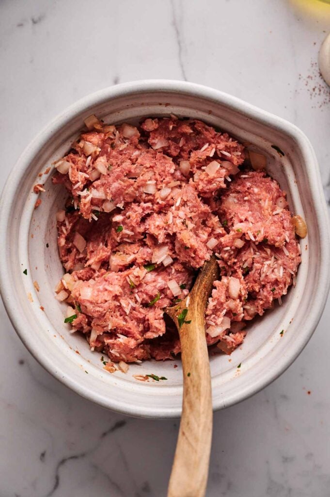 A bowl of raw ground meat mixed with chopped onions and herbs, perfect for preparing delicious Stuffed Cabbage Rolls, with a wooden spoon resting inside the mixture.