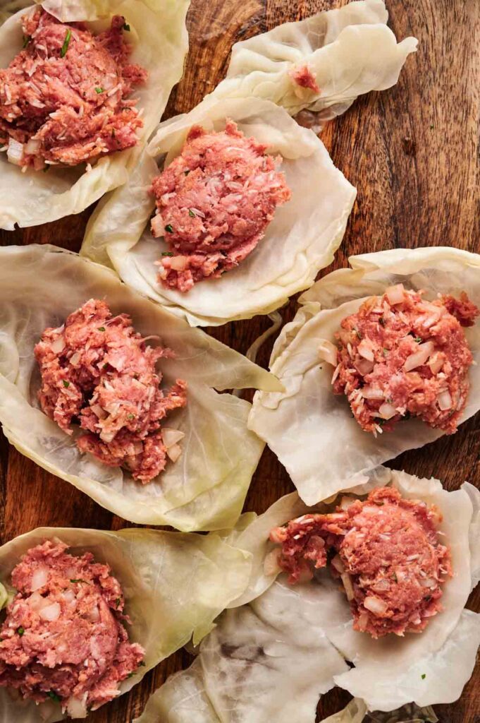 Raw ground meat and chopped vegetables placed on boiled cabbage leaves, arranged on a wooden surface, ready to be rolled into delicious Stuffed Cabbage Rolls.