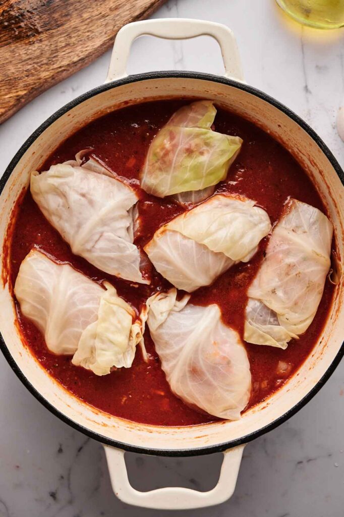 Stuffed Cabbage Rolls simmer gently in a rich red tomato sauce inside a white Dutch oven, creating a comforting scene on the marble countertop.