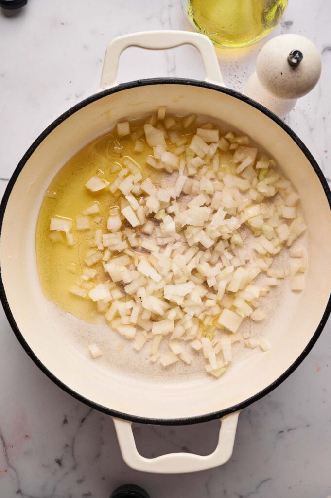 Chopped onions cooking in oil for Stuffed Cabbage Rolls in a white pot on a marble countertop, with a pepper grinder and bottle of oil nearby.