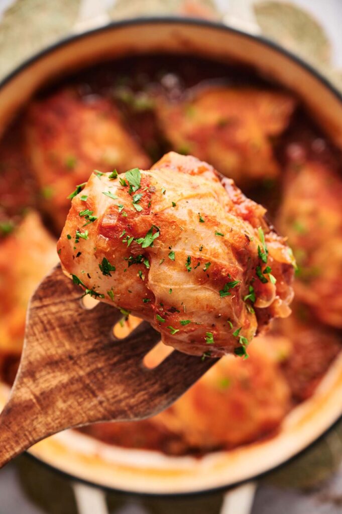 A close-up of a wooden utensil holding a piece of cooked chicken garnished with herbs, with more pieces and stuffed cabbage rolls visible in the pot below.