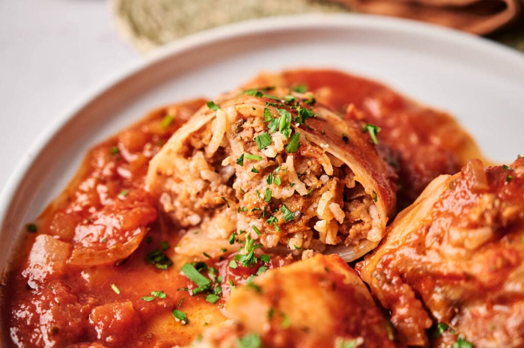 A close-up of Stuffed Cabbage Rolls cut open on a white plate, filled with rice and meat, then topped with tomato sauce and chopped herbs.