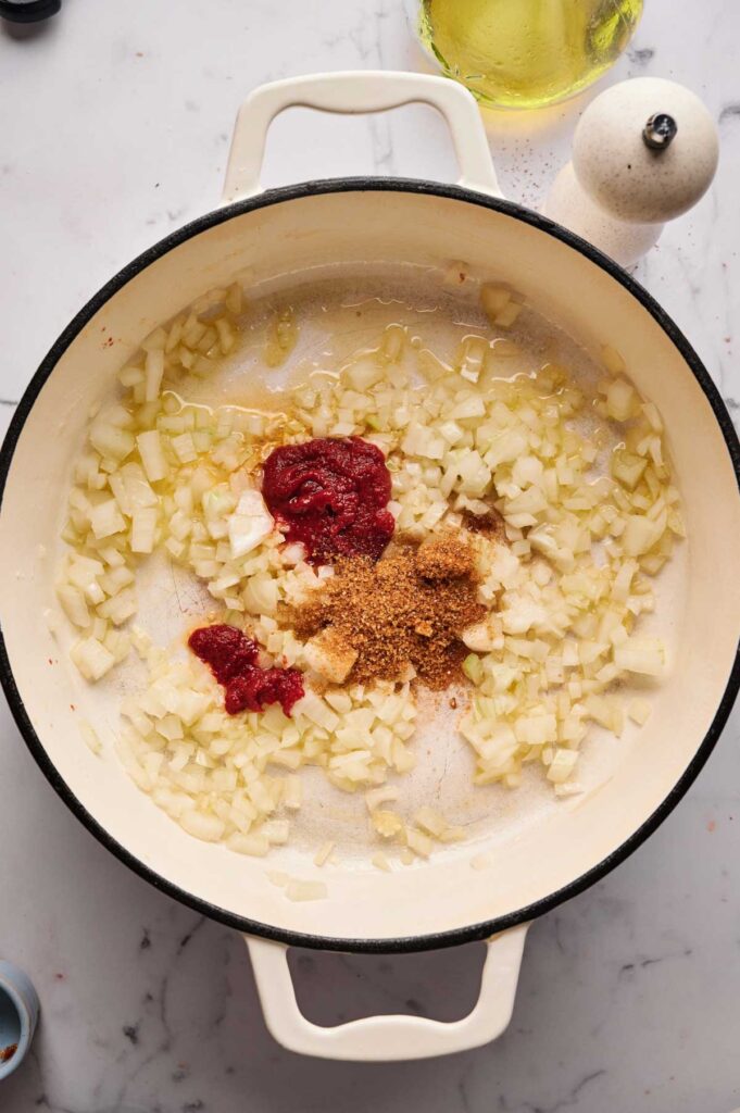 Chopped onions, tomato paste, brown sugar, and spices are cooking in a white Dutch oven on a marble countertop, creating a rich base perfect for Stuffed Cabbage Rolls.