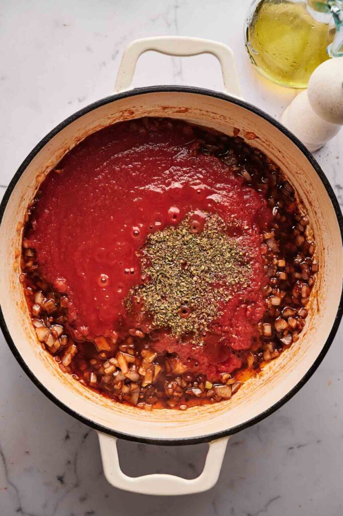 A white Dutch oven containing sautéed onions, tomato sauce, and dried herbs—perfect for making Stuffed Cabbage Rolls—sits on a marble countertop next to a bottle of oil and a pepper grinder.